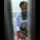 A black girl is video-recorded by her friend sitting on a toilet in a public restroom. She may be peeing or pooping. There are no sounds to indicate which.